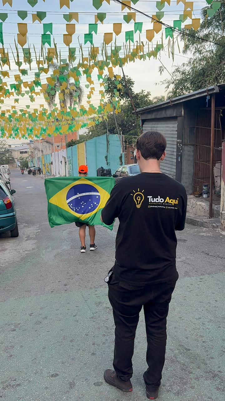 Integrante da Tudo Aqui, de camisa da agência, acompanhando uma gravação na rua com bandeirinhas e uma bandeira do Brasil